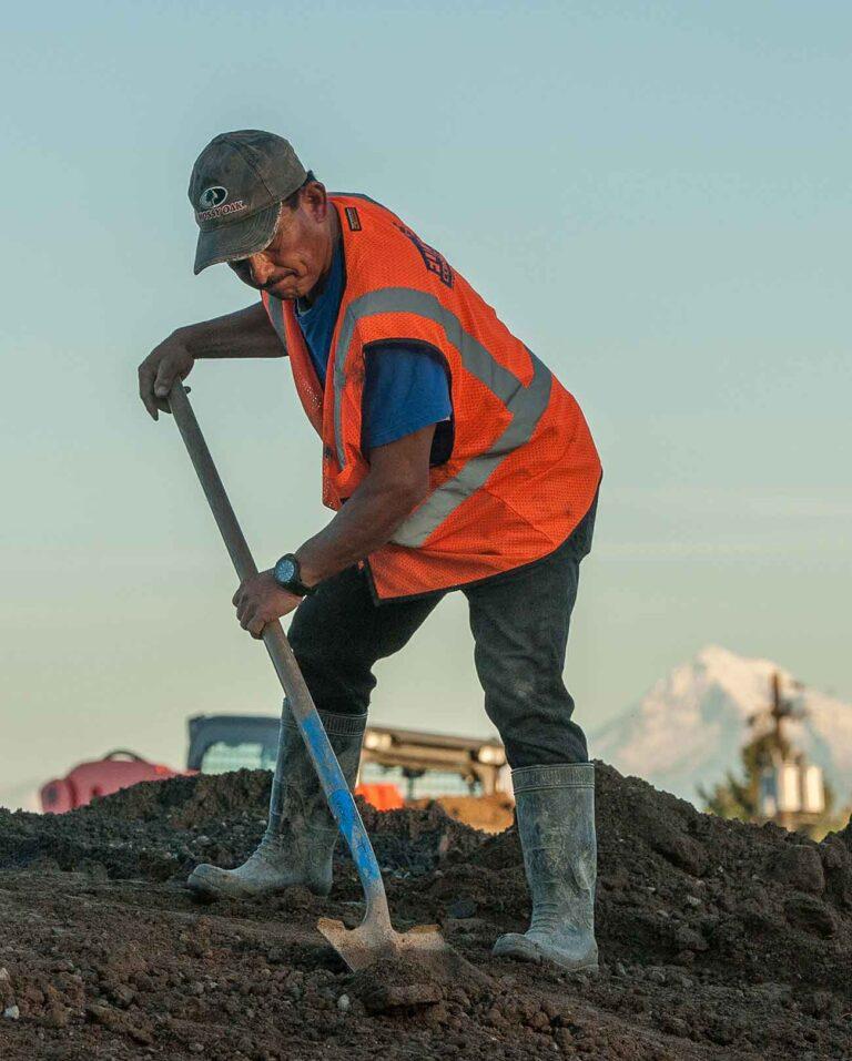 A photo of an adult person working outdoors. They are wearing a baseball cap, work boots, and an orange safety vest. They are shoveling dirt.