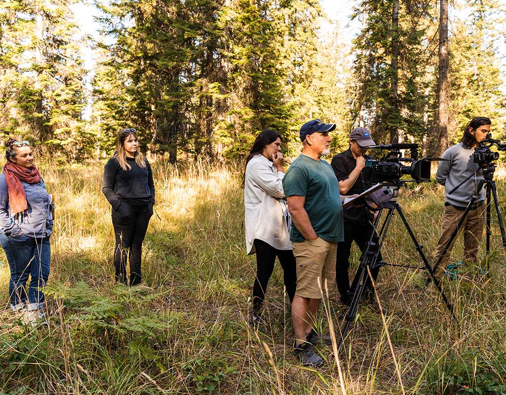 Six individuals stand in a grassy portion of a forest with trees in the background. Two individuals are operating two large film cameras.