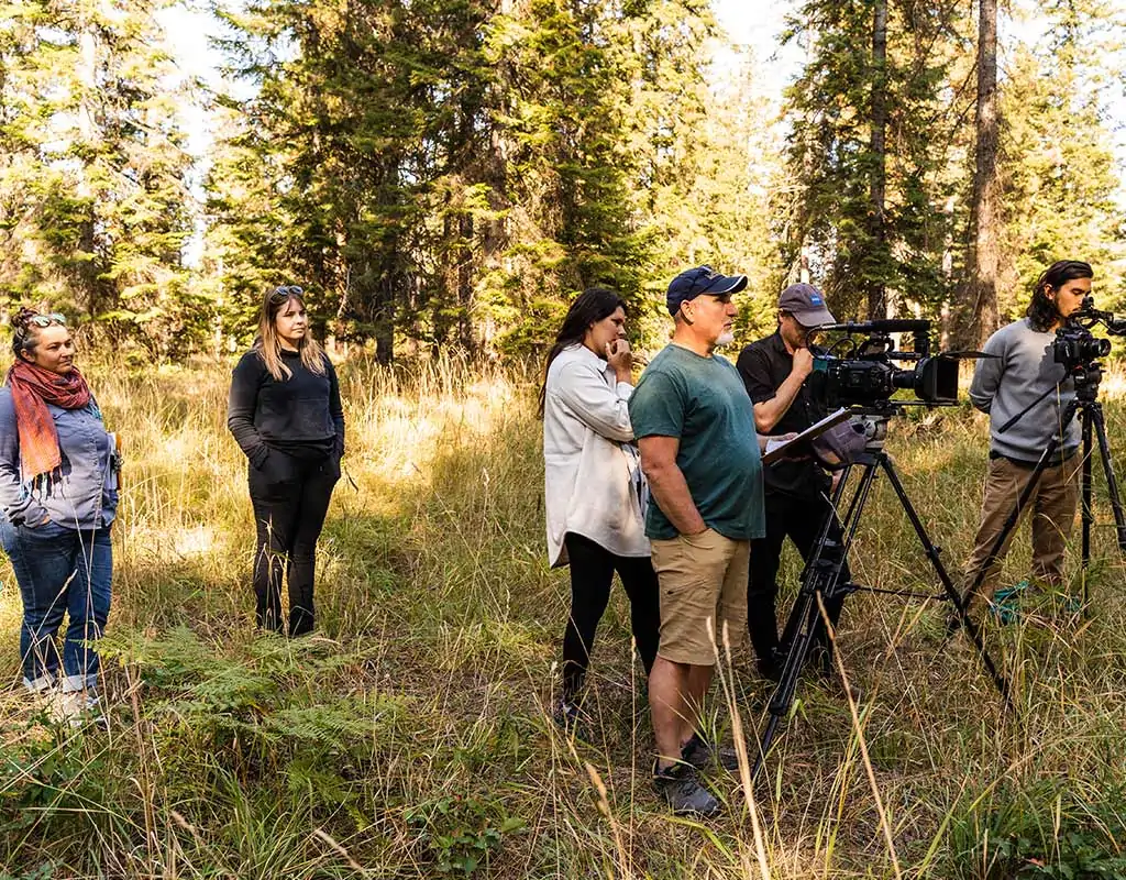 Six individuals stand in a grassy portion of a forest with trees in the background. Two individuals are operating two large film cameras.