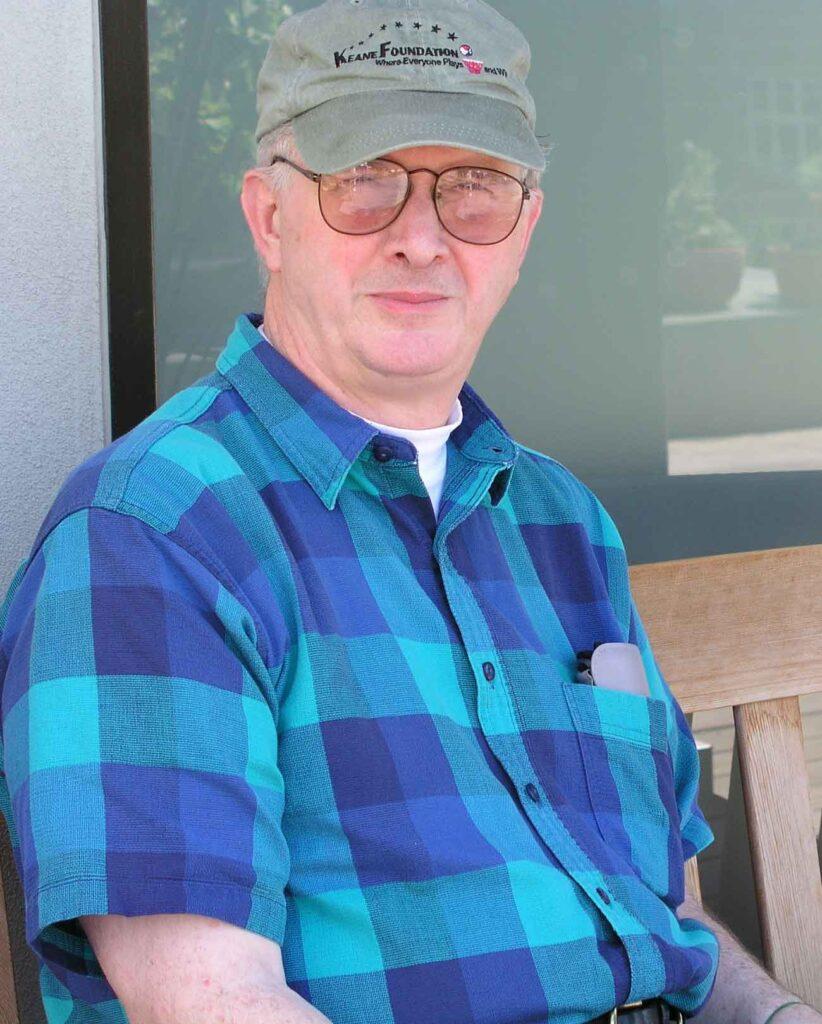 A photo of a middle-aged person gazing at the camera. He is wearing a sage green baseball cap, glasses, and a blue plaid button-up shirt.