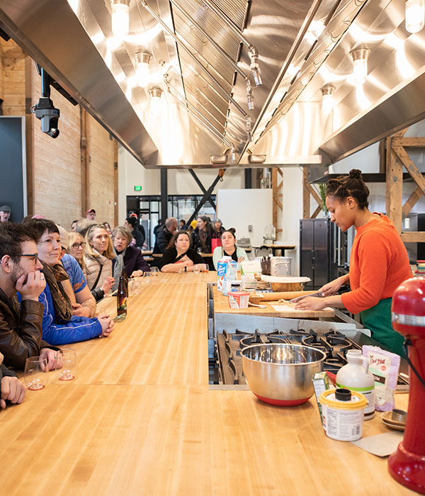 A brand new kitchen counter with a person presenting a cooking demo one one side, and a dozen adults seated on the other side to watch