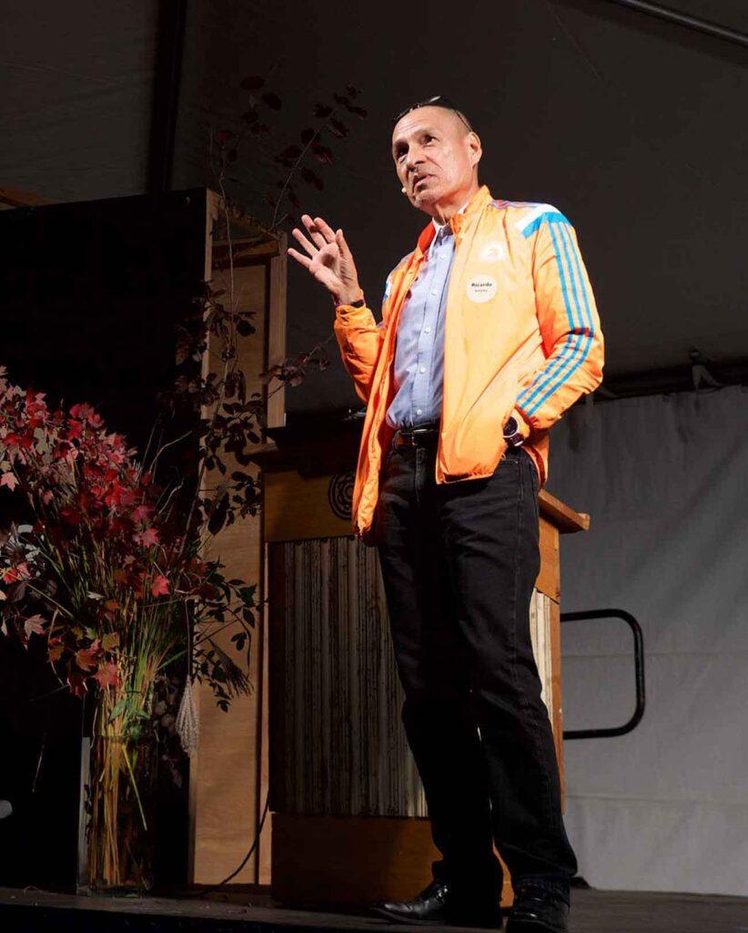 Man in orange jacket stands on a stage delivering a speech on food justice