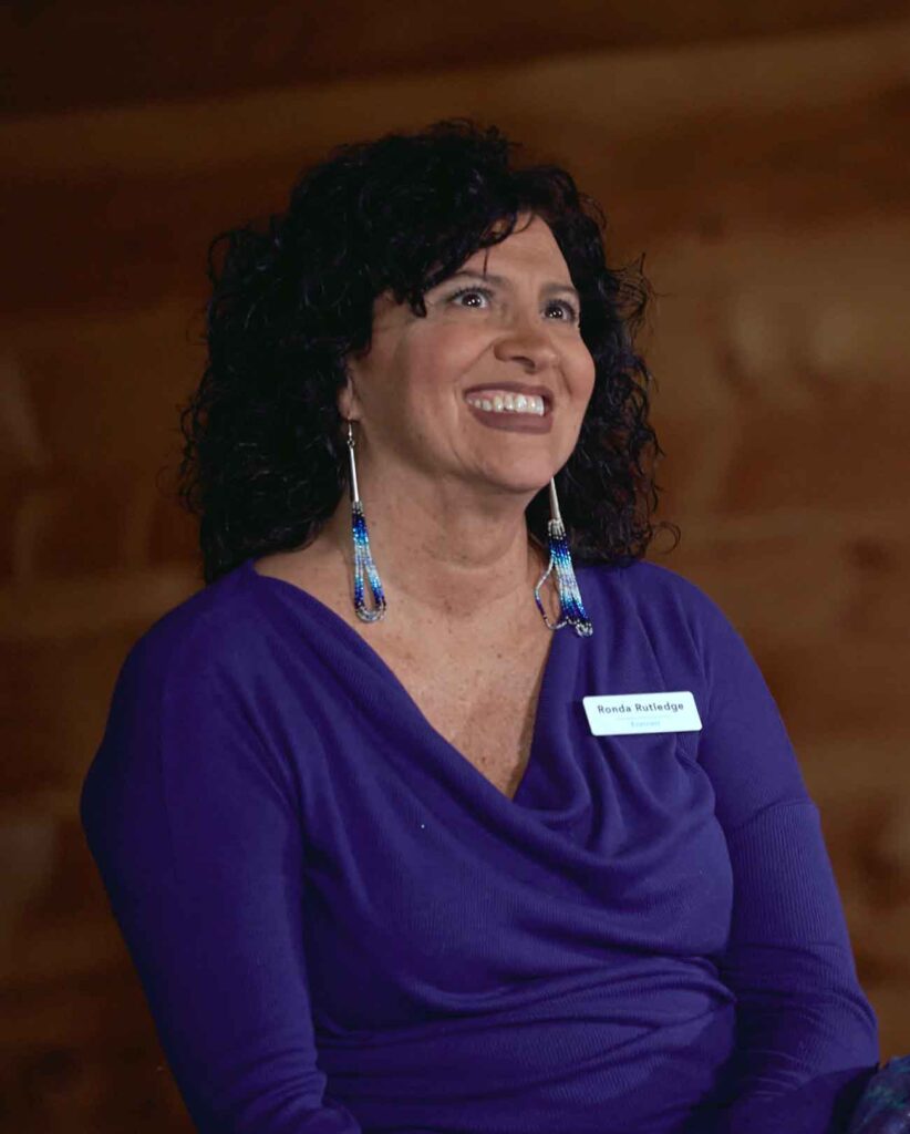 A woman with shoulder-length dark curly hair, wearing long beaded earrings and a purple shirt, smiles while gazing slightly upward at someone on a stage (out of frame)