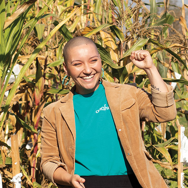 Person standing in front of tall dry corn grinning, fist raised, turquoise shirt and tan blazer