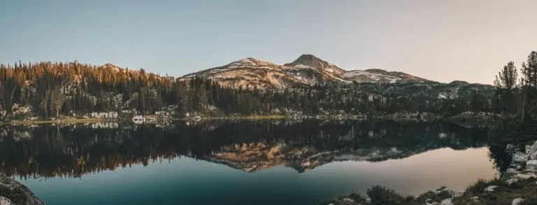 Lake, far shore, and rocky mountain with snow patches and an ombre sky. Mountain and sky also reflected in the still lake water